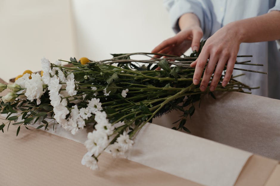 A person with curly brown hair, wearing a dark brown sweater, is arranging a bouquet of white and cream flowers with yellow centers, including daisies and chrysanthemums, in a tall, clear glass vase f