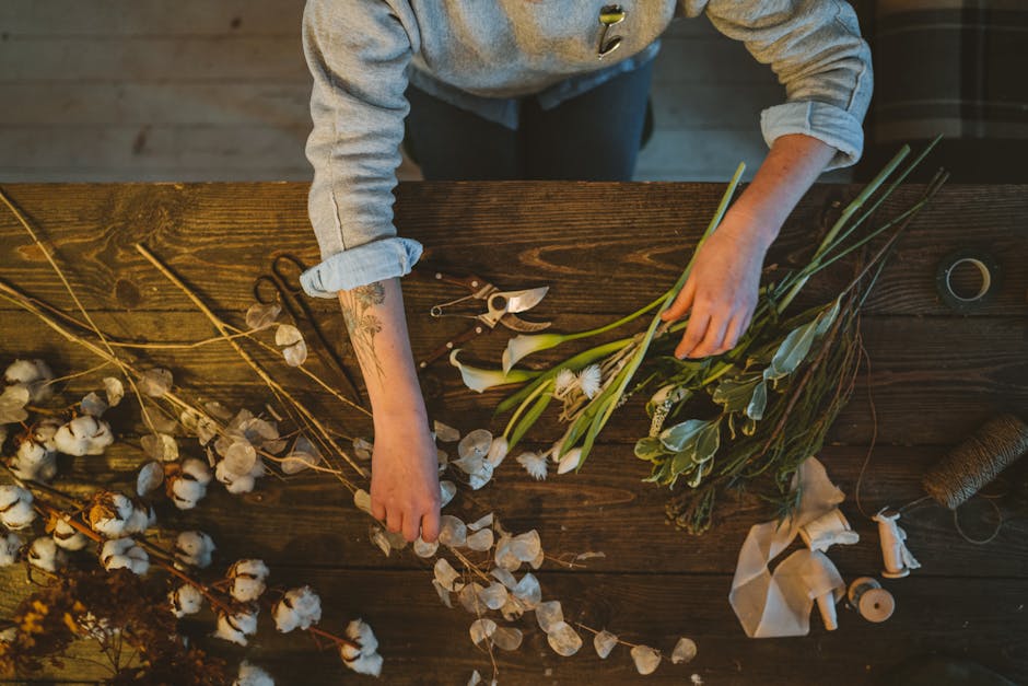 Close-up of a florist's hands arranging a floral bouquet with white, blush pink, and soft green flowers, including roses and delphiniums, wrapped in green paper with ribbons. The arrangement is being 