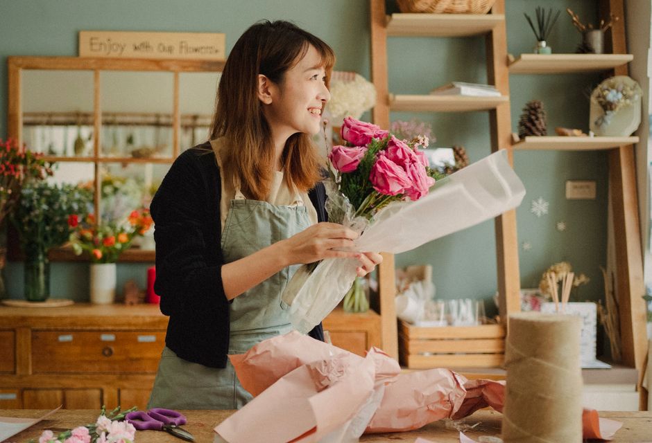 A person holding a fresh bouquet of vibrant yellow sunflowers with large, dark centers and slightly ruffled petals, wrapped in light brown craft paper and tied with a green ribbon. The individual is w