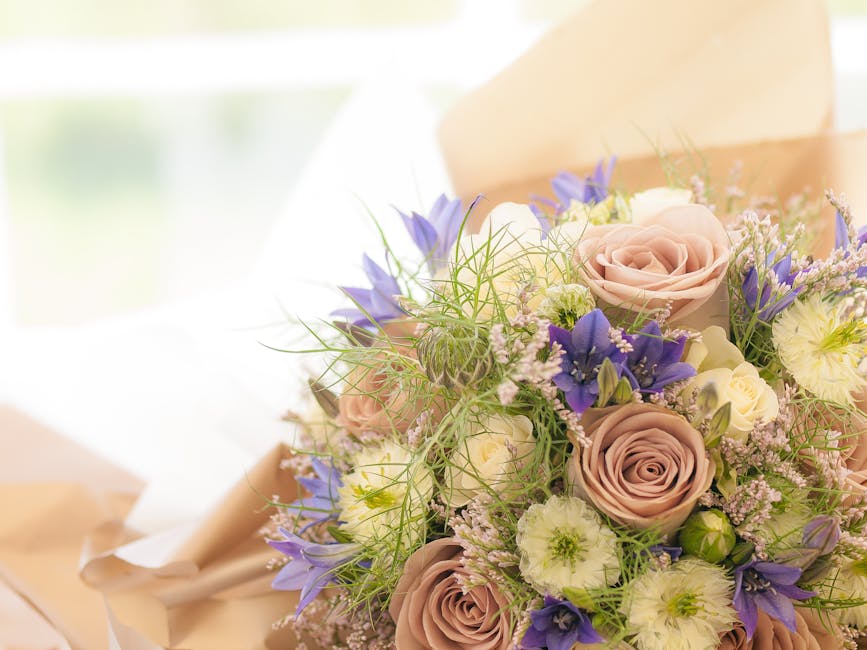A woman with dark, curly hair wearing a light blue face mask and black attire, holding a bouquet of yellow roses tied with a pink ribbon in one hand and a balloon with a 'Happy Birthday' message in co