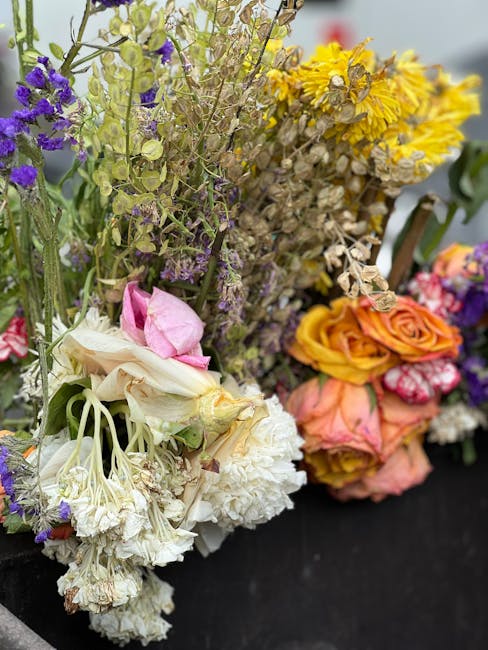 A close-up of a mixed floral bouquet featuring various fresh flowers including white carnations, pale pink roses, purple statice, yellow daisies, and pink-streaked carnations, arranged with green foli