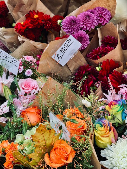 A male delivery person dressed in a red cap and red jacket is holding a floral gift wrapped in colorful geometric-patterned paper with shades of yellow, blue, orange, and pink. The bouquet includes wh